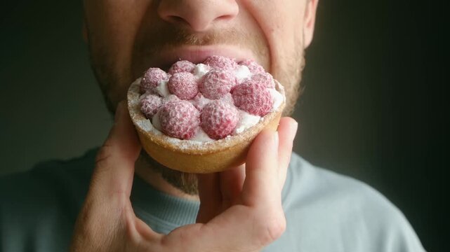 Close up man mouth greedily eating fresh raspberry tartlet. Savors sweet dessert with great pleasure biting in creamy filling fruity topping with gusto. Man eating delicious raspberry tart with cream