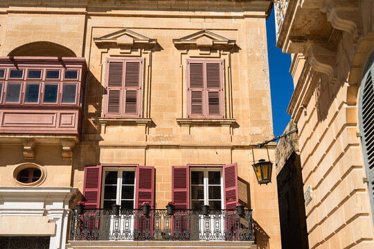 View of traditional limestone building facade with red wooden shutters, a wrought iron balcony, and a classic wooden gallarija under a clear blue sky in Valletta, Port Region, Malta.