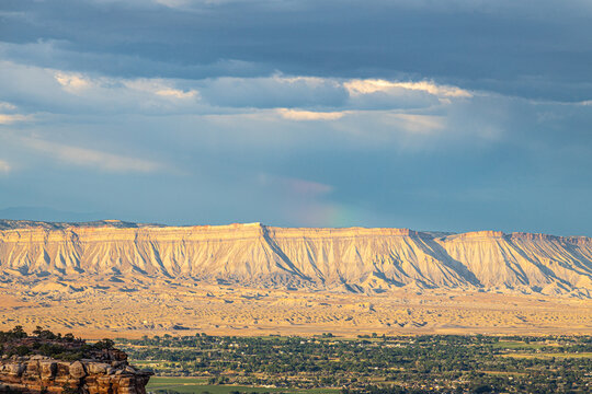 Golden sunlight illuminates the Book Cliffs while rain showers and dark clouds drift across western Colorado.