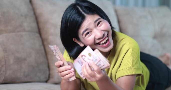 Happy Asian woman counting cash money lying on sofa at home, Thai baht banknote, money, cash, financial success, savings, income, bonus, wealth, and economic stability with cheerful emotion