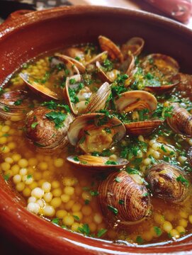 Saffron-infused Sardinian fregola pasta with clams served in a herb broth in a terracotta bowl, evoking a coastal Mediterranean ambiance.