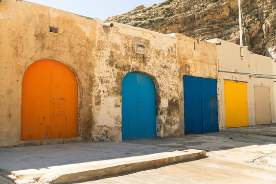 View of traditional boathouses with vibrant colorful doors built into the limestone cliffs at Dwejra Bay in Zebbug, Gozo Region, Malta.