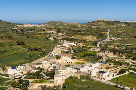 View of traditional limestone houses and lush green fields under a clear blue sky in the rural landscape of Zebbug, Gozo Region, Malta.