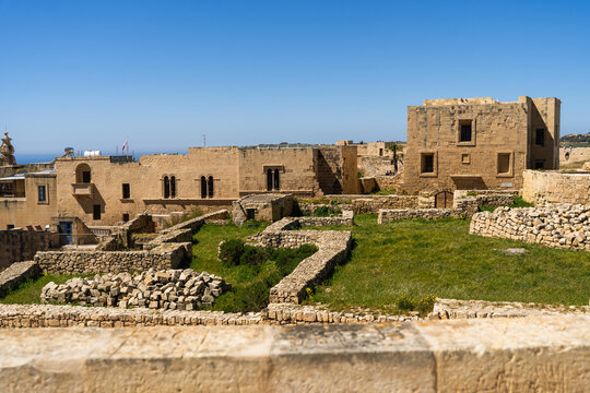 View of ancient limestone buildings and stone ruins with green grass under a clear blue sky in Zebbug, Gozo Region, Malta.