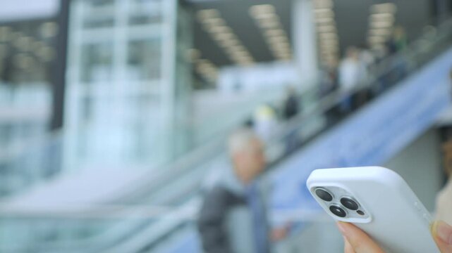 Hand holds smartphone on moving escalator. Background shows blurred indoor space with people. Focused shot on phone with three cameras. Blurred background features glass walls and lights