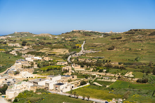 View of traditional limestone houses and terraced agricultural fields across the rolling green hills under a clear blue sky in Zebbug, Gozo Region, Malta.