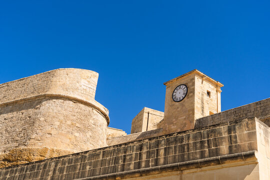 View of historic limestone fortifications and a clock tower under a clear blue sky in Zebbug, Gozo Region, Malta.