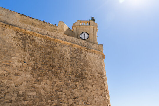 View of the massive limestone walls and a clock tower of a historic fortification under a clear blue sky in Zebbug, Gozo Region, Malta.