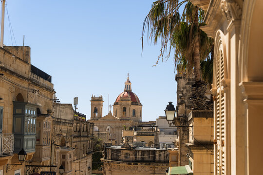 View of the Assumption of the Blessed Virgin Mary Parish Church dome and bell towers rising above traditional limestone buildings and balconies in Zebbug, Gozo Region, Malta.