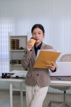 young Asian businesswoman lawyer reading legal document holding coffee cup reviewing contract paperwork standing office workplace professional corporate