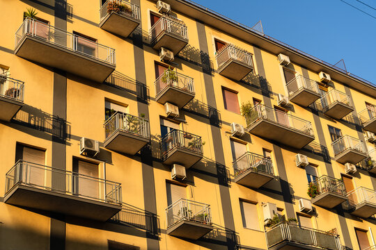 View of the yellow facade of a residential building with numerous balconies and air conditioning units under a clear blue sky in Catania, Sicily, Italy.