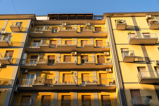 View of a yellow apartment building facade with balconies and shutters under a clear blue sky in Catania, Sicily, Italy.