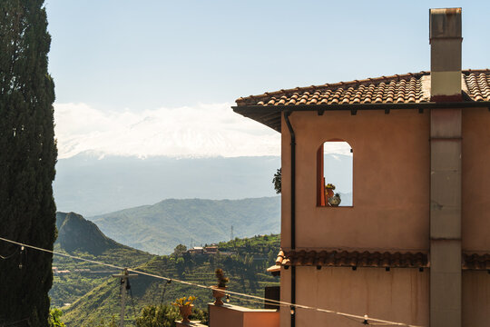 View of a traditional building with a terracotta roof and chimney overlooking the snow-capped Mount Etna and green hills in Castelmola, Sicily, Italy.