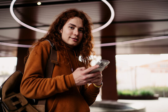 Smiling man with curly hair looking at camera holding smartphone, male freelancer using mobile banking app outdoors, authentic representation of young adult with cellphone, 5G data, Caucasian model.