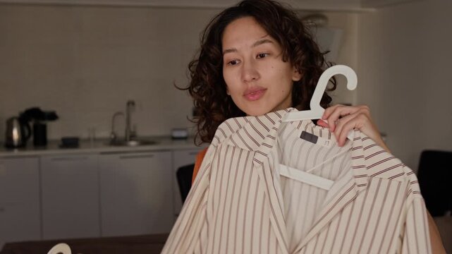 Asian young adult woman comparing striped and patterned shirts on hangers at home. Clip is ideal for fashion vlogging, wardrobe styling, and retail marketing