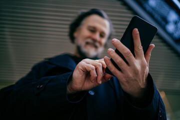 Fototapeta na wymiar Close up of senior man hands using smartphone with blurred background, mature professional male interacting with mobile app on touchscreen device in modern environment, copy space.