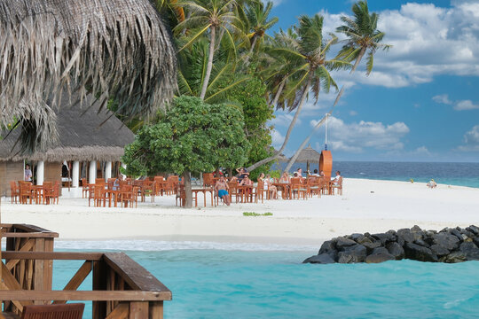 View of people dining at the Fihalholi beach resort restaurant with wooden tables on white sand under palm trees and a thatched roof in Kaafu Atoll, Kaafu Atoll, Maldives.