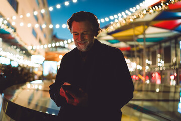 Fototapeta premium Smiling businessman using mobile phone app, night city street festive background, caucasian middle aged model, browsing content online, digital nomad working remotely, high speed 5G internet.