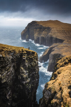View of the dramatic Asmundarstakkur sea stack and towering cliffs with a person standing on a precipice under a moody sky in the Faroe Islands.