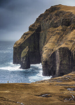 View of the dramatic sea stack Asmundarstakkur and towering cliffs with a person in a yellow jacket on a grassy plateau overlooking the Atlantic Ocean in the Faroe Islands.