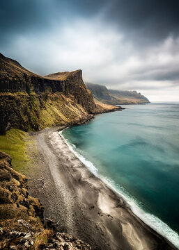 View of the dramatic coastline with steep green cliffs and a black sand beach meeting the turquoise ocean under a moody sky Vagar, Faroe Islands.