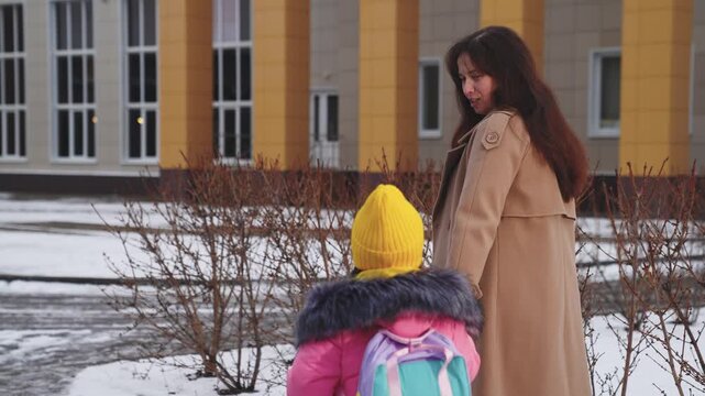 happy family, winter school yard, schoolgirl with her mother walks through school yard by hand, concept happy family, little schoolgirl walks with her mother through school yard with backpack, mother