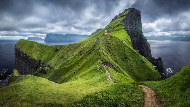 View of Kallur Lighthouse on a steep green cliff with a narrow dirt path and dramatic clouds over the ocean Klaksvik, Northern Isles, Faroe Islands.