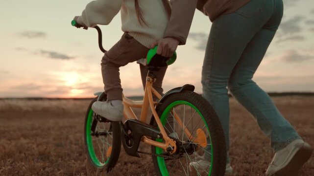 Teaching independence to kids, Field biking session, Cherished moments at twilight, Outdoor family leisure, Support in learning new skills, Bonding through challenges, Achieving new milestones, Mother
