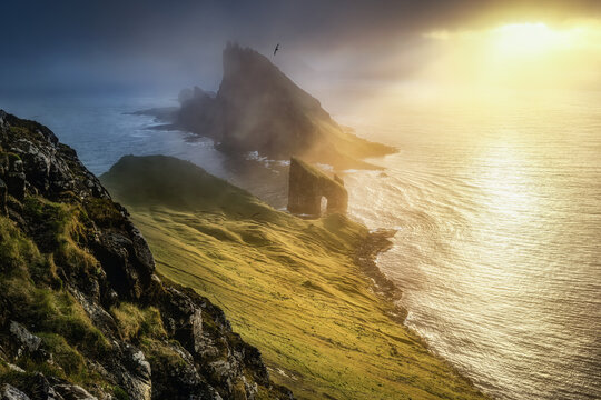 View of the Drangarnir sea stacks and Tindholmur islet amidst misty ocean waves and green cliffs during a golden sunset Faroe Islands.