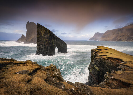 View of the Drangarnir sea stack with crashing waves and rugged cliffs under a dramatic sky in Faroe Islands.