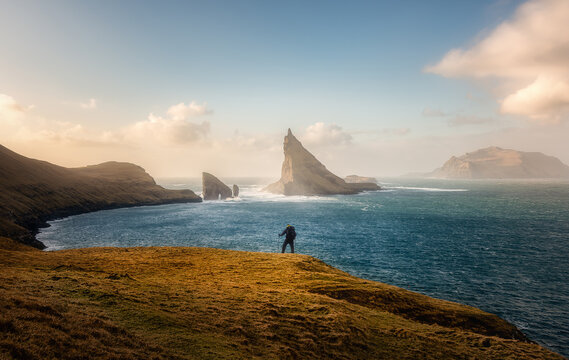 View of a hiker standing on a grassy cliff overlooking the iconic Drangarnir sea stacks and Tindholmur island under a soft golden sky in Faroe Islands.