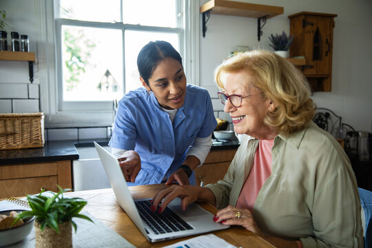 Caregiver helping senior woman use laptop in home kitchen