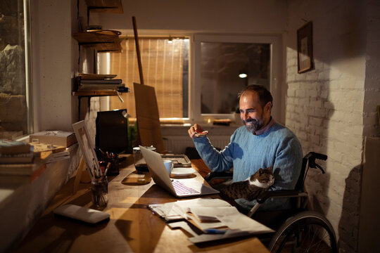 Smiling man in wheelchair working on laptop at home office with cat at night