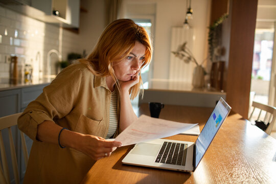 Concerned woman reviewing bills at laptop in home kitchen