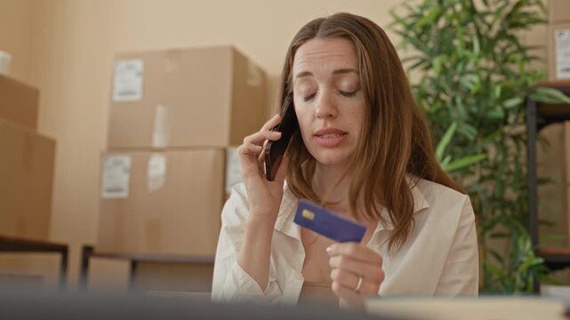 Woman holding creditcard and smartphone to ear while sorting parcels amid stacked boxes in a small business packing area building; order processing focused.
