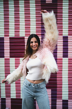Joyful woman raising one arm in celebration against colorful striped background, happy girl in stylish faux fur coat expressing freedom and success, emotional connectivity and urban lifestyle.