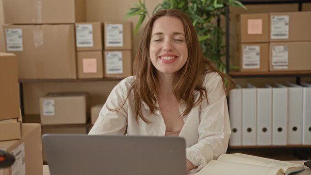 Woman sitting at desk typing on laptop amid stacked shipping boxes and binders in a packing station inside a building; small business confidence.