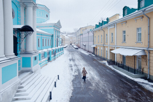 View of a person in a brown coat walking on a snow-covered street lined with colorful blue and yellow historic buildings under an overcast winter sky in Moscow, Moscow, Russia.