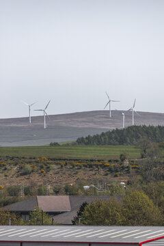 View of wind turbines standing on a rolling hill above green fields and industrial rooftops under a clear sky Lancaster, England, United Kingdom.