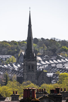 View of the tall stone spire of a Gothic church rising above rows of terraced houses and green trees under a clear sky in Lancaster, England, United Kingdom.