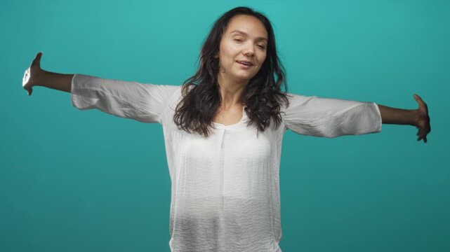 Woman stretching arms wide with eyes closed and slight smile, wearing white blouse in a teal studio setting; calm reflection.