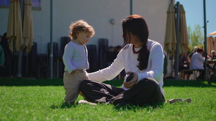Fototapeta premium Mother placing baseball cap on toddler during playful outdoor family moment on sunny grass lawn conveying affection parenting warmth and joyful childhood connection