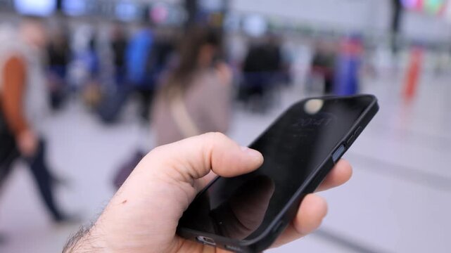 Hand holds smartphone in public space. Blurred background shows moving people. Person focuses on device, calm and intent. Footage suits travel, tech, or communication themes