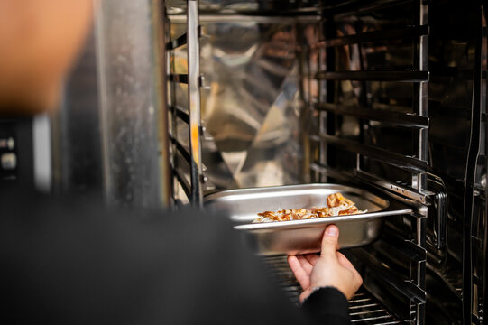 A chef slides a stainless steel tray filled with cooked food into a professional commercial combi oven in a restaurant kitchen.
