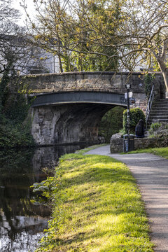View of a historic stone bridge over the Lancaster Canal with a person sitting on the towpath and lush greenery Lancaster, England, United Kingdom.