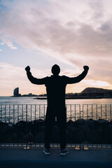 Fototapeta premium Athlete standing on modern bridge at sunset, man in black sportswear celebrating success with raised arms, triumph and achievement concept, urban wellness and fitness motivation, harbor view.