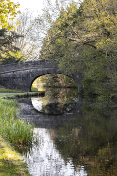 View of a historic stone arch bridge reflecting in the calm waters of a canal surrounded by lush green trees and spring foliage in Lancaster, England, United Kingdom.