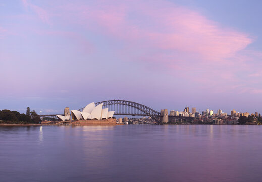 View of the Sydney Opera House and Sydney Harbour Bridge under a pink sunset sky with calm water reflections in Sydney, New South Wales, Australia.