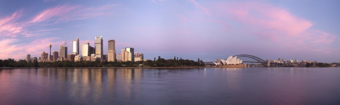 View of the Sydney Opera House and Harbour Bridge with the city skyline under a pink sunset sky reflected in the water Sydney, New South Wales, Australia.