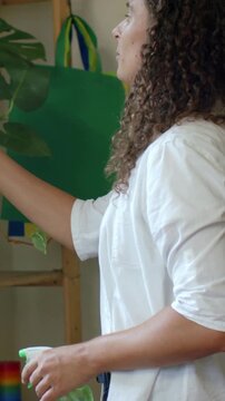 Vertical video: Misting adult in white shirt with green spray tidying pothos on shelf, copy space